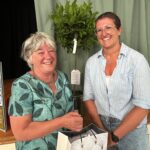 Kerry Bartlett and Yvonne Gallimore pose together for a photo, they both have big laughing smiles. There is a small tree behind them and Yvonne is holding a gift bag.