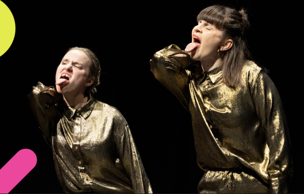 2 female performers dressed in shiny gold shirts and trousers are attempting to lick their elbows.