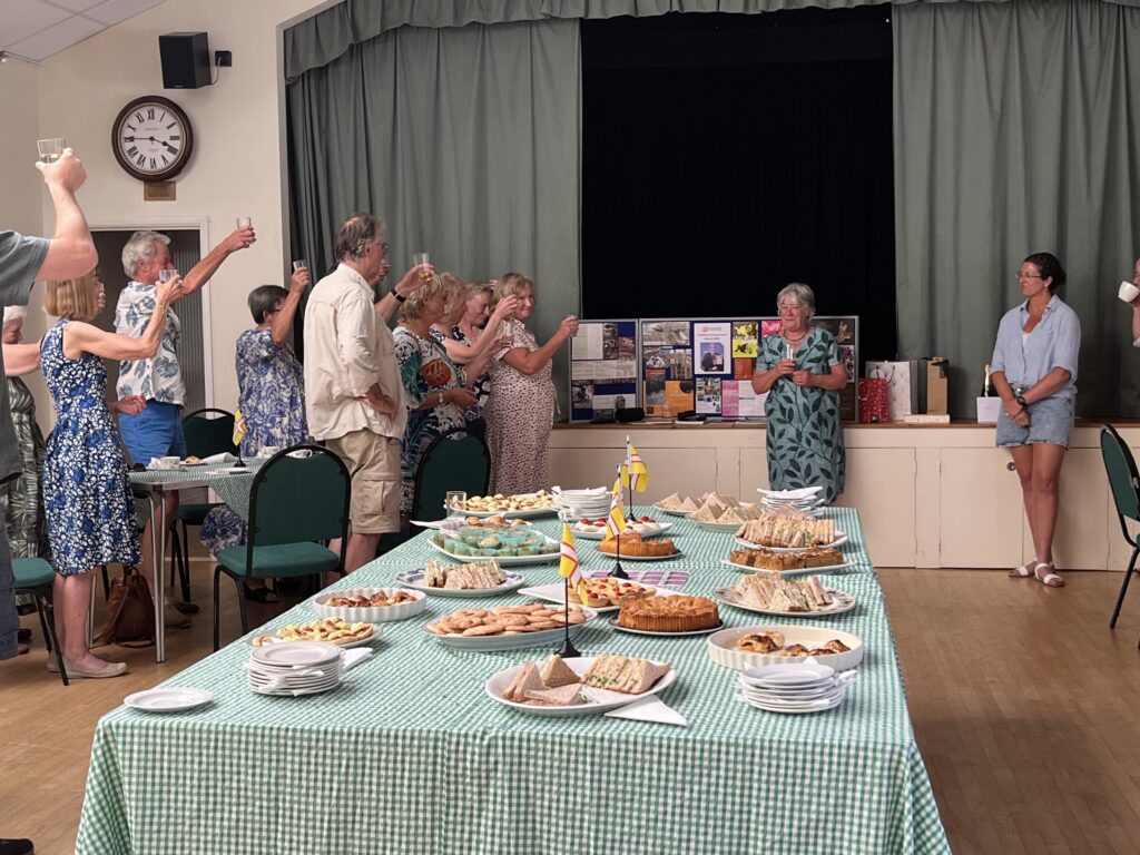 Yvonne's retirement party - a group of people are raising their glasses in a toast to a woman standing in the centre. There is a big table of buffet food in the front.
