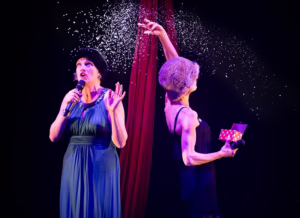 Two performers, one facing front in a blue dress, looking up at snow falling. The other is turned away and is throwing fake snow behind them.