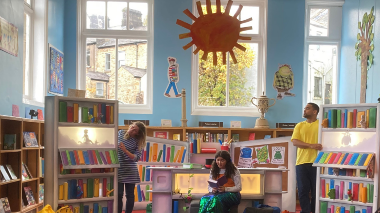 Three performers in a colourful children's library, surrounded by illustrated bookshelves and playful decorations. One performer sits reading a book, while two others stand beside large theatrical bookshelves featuring illuminated silhouettes.