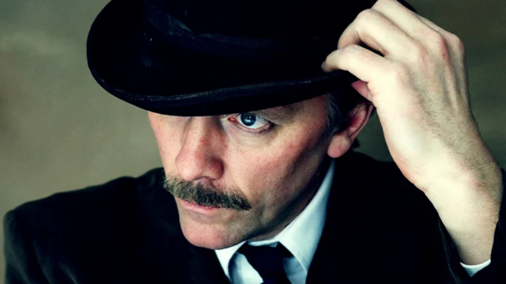 Rural Touring Lead Image: Close-up portrait of a man with a dark moustache, wearing a black suit, white shirt, black tie and bowler hat. He lifts the brim of his hat with one hand, revealing one blue eye looking directly at the camera. The image has dramatic lighting and a muted green-grey background.