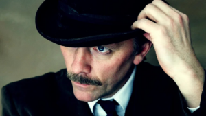 Rural Touring Lead Image: Close-up portrait of a man with a dark moustache, wearing a black suit, white shirt, black tie and bowler hat. He lifts the brim of his hat with one hand, revealing one blue eye looking directly at the camera. The image has dramatic lighting and a muted green-grey background.